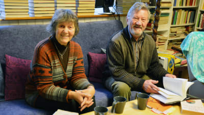 Siv Långstedt och Torsten Bergman sitter i en blå soffa, omringad av böcker. På bordet står böcker och tre kaffekoppar.