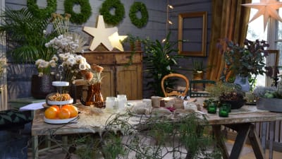 En del av ett rum som är pyntat i gröna och vita juldekorationer. På bordet finns olika pysselmaterial som torkade blommor, torkade apelsinskivor, olika juteband och snören, torkat gräs.