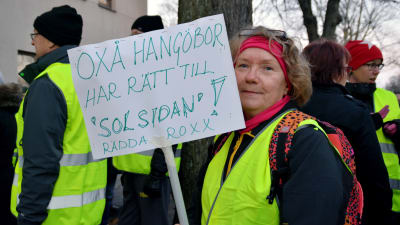 En kvinna står i en gul reflexväst. I handen håller hon en skylt där det står "Oxå Hangöbor har rätt till "solsidan". Rädda Roxx!".