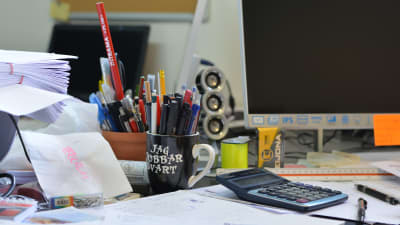 ett skrivbord fyllt med pennor, ritningar, dator och en svart mugg med texten "jag jobbar svart".