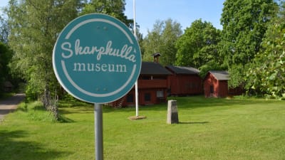 En rund skylt i turkosblått och vitt. Det står Skarpkulla museum på skylten. Bakom finns lador i rött. Sommar och grönt gräs.