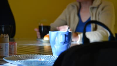 En kaffemugg och smulor på ett fat. I bakgrunden sitter en person och njuter av kaffe och bulla.