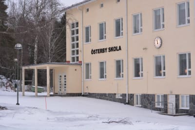 En skolbyggnad i flera våningar byggd i ljusmålad betong. Det står Österby skola på fasaden. På väggen finns också en stor, rund klocka. Vinter och snö, inga skolbarn.