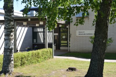 Ingång till vit tegelbyggnad. Träd och gräsmatta utanför.