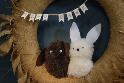 två kaniner gjorda av garnbollar sitter i en krans med textvimpel där det står "glad påsk"