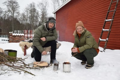 En man och en kvinna som står på huk och eldar i friluftskök gjorda av konservburkar, utomhus på vintern.