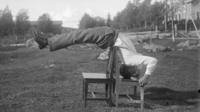 En man leker leken "bryta rygg", där han ställer sig på huvudet på en stol och lutar kroppen bakåt över en annan stol. Bilden är tagen 1931 i Närpes, Kalaks.