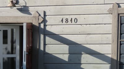 Fasaden på ett gammalt trähus i beigegrön färg. Siffran/årtalet 1810 på väggen. Solsken.