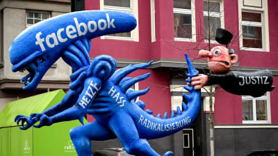 Sinisen alien patsaan hännästä pitää kiinni lennossa oleva tuomarihahmo. Alien edustaa Facebookkia ja tuomari oikeutta.