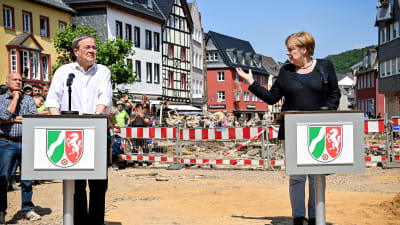 Till höger Angela Merkel och till vänster Armin Laschet. Bakom dem rödvit avspärrning och längst bort i bakgrunden en rad med hus.