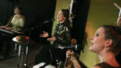 Tre musiker i festklänningar sitter på en musikscen.  
