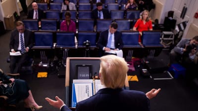 Donald Trump sedd bakifrån i pressrummets talarstol sträcker ut händerna.