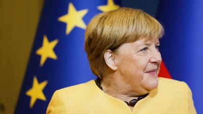 En bild på Tysklands förbundskansler Angela Merkel. Hennes blick är riktad bort från kameran. I bakgrunden ser man EU:s flagga. 