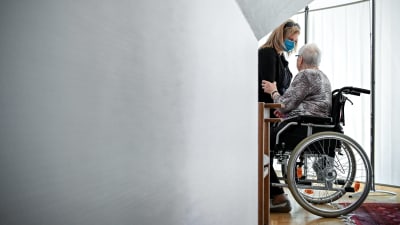 En person i munskydd hjälper en äldre person i rullstol.