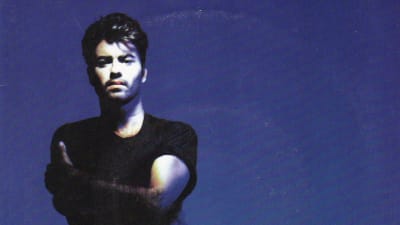 Nuori George Michael seisoo sinisen taustan edessä kädet puuskassa ja katsoo suoraan kameraan. 