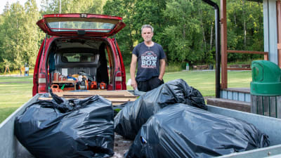 En man står vid en röd paketbil med en släpvagn. I släpvagnen finns stora svarta plastsäckar.