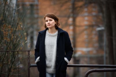 Iiris Viljanen utomhus, i stickad tröja och yllekappa, med husvägg i tegel bakom.