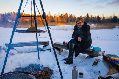 En kvinna i vinterkläder sitter utomhus vid en grillplats