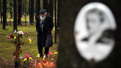 Gammal skäggig rysk man betraktar en släktingsgravkors i skogen. I förgrunden på ett träd ett oskarpt foto på en kvinna..