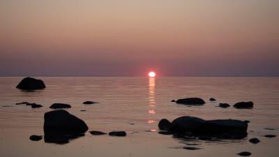 Aurinko on laskemassa horisonttiin kivikkoisella merenrannalla.