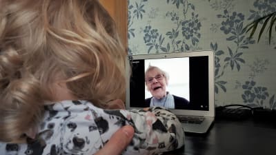 Ett barn med lockigt hår ser på en dataskärm med en bild på en äldre kvinna som ler. 