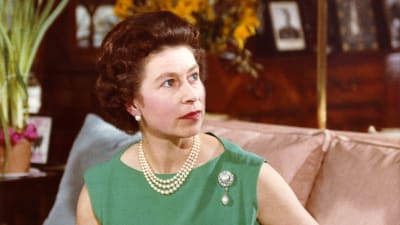 Drottning Elizabeth II i grön klänning sitter på en soffa med en av sina hundar, en welch corgi pembroke, i famnen.