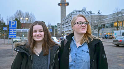 Helmi Nurkkala tittar in i kameran, Ida Salo tittar ut ur bilden, då de poserar på en parkeringsplats med Yletornet i bakgrunden.