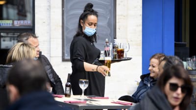 En kvinnlig servitör håller i en bricka med öl på en uteservering. Hon har munskydd på sig.