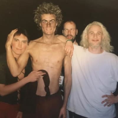 Neljä nuorta miestä teatterimaskeissaan poseeraavat kaulakkain: toisena vasemmalta nuori Mikko Kanninen ilman paitaa, kiharainen tukka pörrössä, kulmakarvat tummiksi ehostettuina.