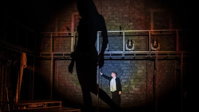 En man står på en teaterscen, en stor mörk skygga bredvid sig.