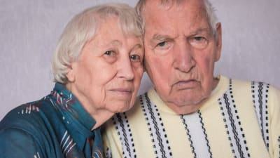 Ett äldre par i blå och gul blus. Det har lutat sina huvuden mot varaandra och ser ledsna eller bekymrade ut i sina ansikten