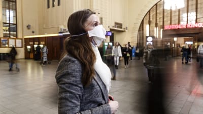 En kvinna står och väntar på någonting på Helsingfors järnvägsstation. Hon har ett vitt munskydd framför munnen och näsan.