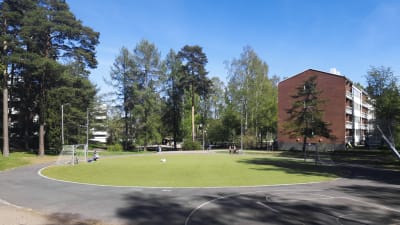 En park med en gräsplätt på mitten. På höger sida syns ett rött tegelhus,