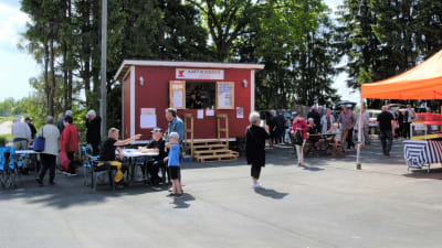 Bilden föreställer ett sommartog på Vessö. På bilden syns ett cafè, samt människor som besöker torget.