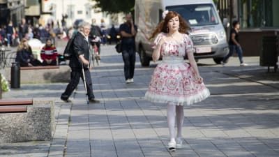En kvinna klädd i så kallade Lolitakläder - en fluffig vit och rosa klänning med spetsar på. Hon har vita högklackade skor och blommor i håret. Hon går på en trottoar i centrum av Vasa. I bakgrunden tittar en äldre kvinna på henne.