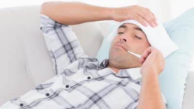 En man ligger på en soffa med en termometer i munnen och en handduk på huvudet.