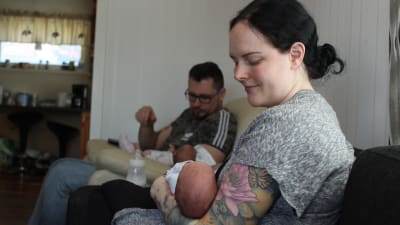 Kvinna som sitter i soffan med ett spädbarn i famnen, i bakgrunden sitter en man med ett annat spädbarn i famnen. 