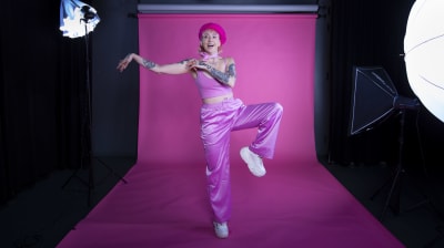 Ronja Stanley står på ett ben i en fotostudio där man ser endel lampor och en rosa vägg, Ronja skrattar.
