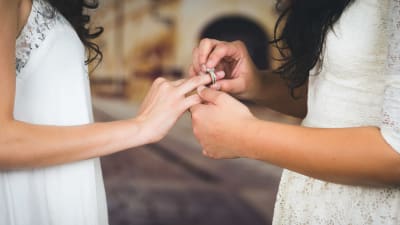 En kvinna i bröllopsklänning sätter en ring på fingret på en annan kvinna i bröllopsklänning.