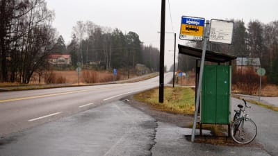 En busshålplats intill en landsväg sedd från sidan. Inga människor syns i bild, en cykel står parkerad bakom busskuren.