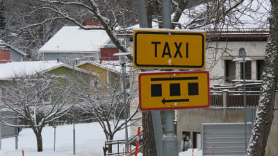 En taxiskylt i gult med svart text och en skylt som visar marlerade parkeringsrutor på en stolpe utanför Raseborgs sjukhus i Ekenäs. Man ser inte sjukhuset men nog en del småhus. Vinter och snö. 