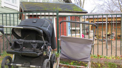 Några barnvagnar framför en ingärdad gård med lekstuga och klätterställning. Man kan skymta några små barn med ryggarna mot kameran.