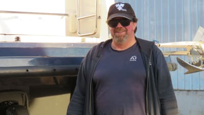 Guy Sannholm, en man med keps och solglasögon står utanför en stor hall bredvid en båt som står på pallar.