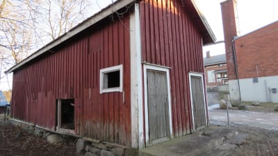 Ett förfallet förråd eller gammalt träskjul som har ruttet virke och flagande färg på fasaden. I bakgrunden ser man Bromarv skola.