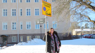 Arbetarskyddsfullmäktig Piia Hasselberg står utanför Raseborgs stadshus, håller i en vägskylt där det står Räddningsväg -pelastustie och en symbolisk bild på en räddningsbil med lyft stege.