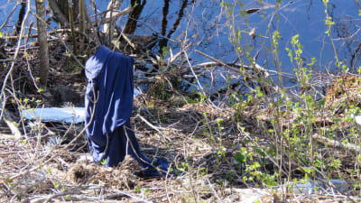 Skräp och en kvarlämnad blå tröja på en rastplats invid en sjö. Vår och sol.