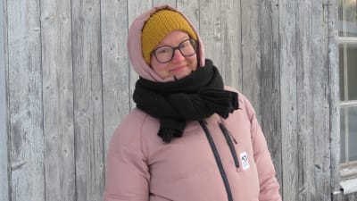 Susanna Kankare i varma vinterkläder i rosa. Huvan uppdragen. Det är vinter och hon står framför en grå trävägg.