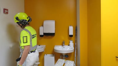 Kvinna i hjälm och gul refexvägg målar väggar vid ett tvättställ i Sjundeås blivande nya skolcentrum. Väggarna går i vitt, gult och orange.