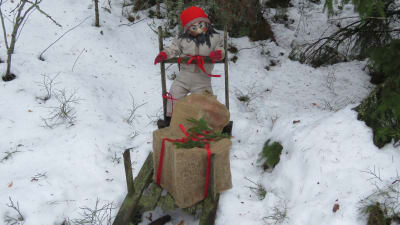 En handgjord tygtomte ute i vinterlandskap. Tomten står bakom en gammaltida kälke. På kälken ett julpaket av juteväv med röda band och grankvist.