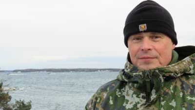 Portättbild på brigadkommendör, kommodor Juha Kilpi, Nylands brigad. Han står ute i Syndalen, vinter. Bakom hav och man kan skymta några Jurmobåtar, öppet vatten.
